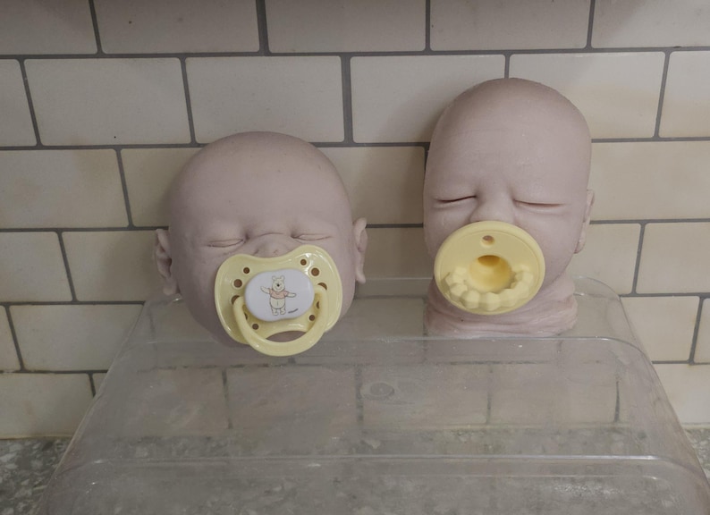 Pu&ograve; includere: Due teste di bambola pallide e calve con gli occhi chiusi. La testa a sinistra presenta un ciuccio giallo chiaro con un'immagine di Winnie the Pooh. La testa a destra presenta un ciuccio giallo chiaro con denti e gengive bianche modellate.