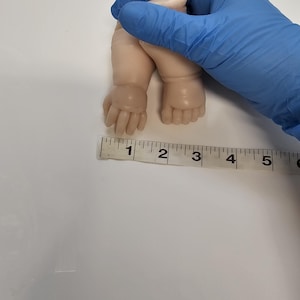 Pu&ograve; includere: Una coppia di arti per bambole in vinile pallido, liscio e non dipinto, composta da due mani e due piedi. Un metro a nastro bianco &egrave; posizionato orizzontalmente sotto gli arti, mostrando una larghezza di circa 11,5 centimetri.