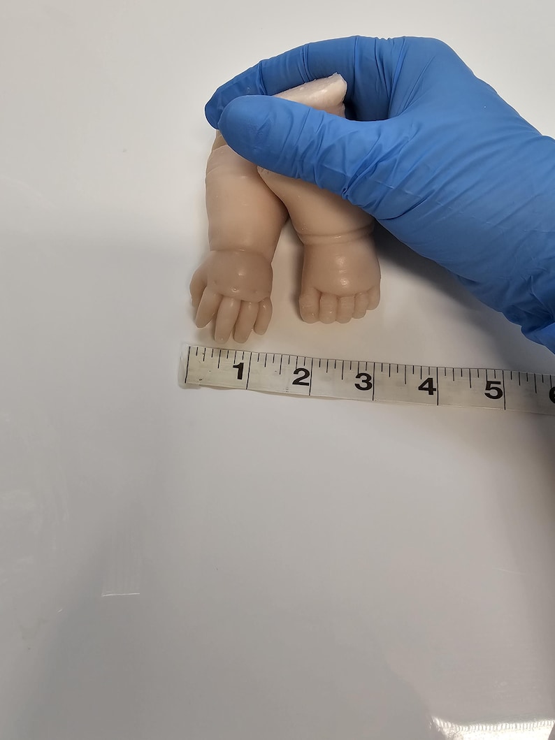 Pu&ograve; includere: Una coppia di arti per bambole in vinile pallido, liscio e non dipinto, composta da due mani e due piedi. Un metro a nastro bianco &egrave; posizionato orizzontalmente sotto gli arti, mostrando una larghezza di circa 11,5 centimetri.
