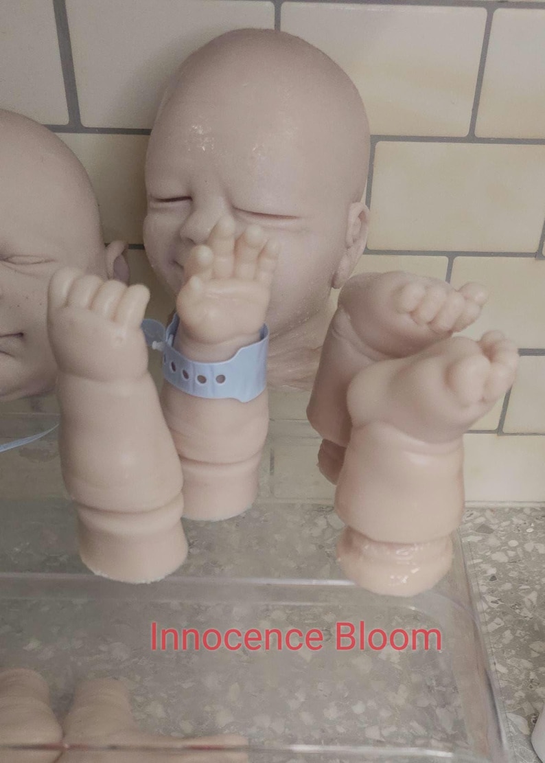 Pu&ograve; includere: Un kit per bambola reborn in vinile dalla pelle chiara composto da una testa con occhi chiusi, due braccia e due gambe. Un braccio indossa un braccialetto identificativo ospedaliero in plastica azzurra. Il testo Innocence Bloom &egrave; stampato in rosso.