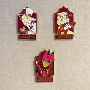 Puede incluir: Tres pines de esmalte con personajes de dibujos animados en marcos burdeos. Uno dice "BIG BOSS ENERGY" y presenta un personaje con sombrero blanco. Otro dice "I BELIEVE IN YOU". El tercer pin dice "JUST SMILE ANYWAY".