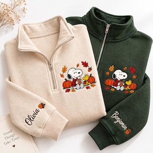 May include: Two quarter-zip sweatshirts, one cream and one dark green, featuring embroidered Snoopy and Woodstock characters surrounded by autumn leaves and pumpkins. The cream sleeve is embroidered with the name Olivia, and the green sleeve with Benjamin.