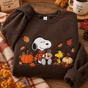 Puede incluir: Una sudadera de cuello redondo en color marrón oscuro con un diseño bordado de Snoopy usando una bufanda y sosteniendo una taza, junto a Woodstock, calabazas, bellotas y hojas de otoño. El gráfico incluye vapor y una huella de pata.