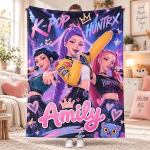 May include: A rectangular fleece blanket featuring a vibrant K-pop themed graphic with three stylized singers holding microphones. The design includes the text K-POP, HUNTRX, and the name Amily in large pink letters, surrounded by stars, hearts, and a crown.