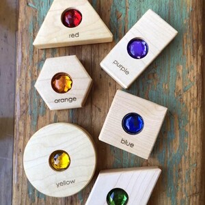 May include: Wooden shapes with colourful gems in the centre. The shapes are a triangle, rectangle, hexagon, circle, and diamond. The gems are red, purple, orange, yellow, blue, and green. The words "red", "purple", "orange", "yellow", "blue", and "green" are written below each gem.