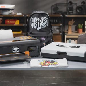 May include: Two black and white desktop printers with panda face graphics, a black backpack with the word Hype, a grey bag with the word Just, and a white t-shirt with colorful graffiti text. The printers feature orange accents and paper rolls.