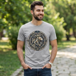 May include: A heather grey short-sleeved t-shirt featuring a circular graphic on the chest. The design depicts a detailed lion head in profile, rendered in shades of gold, brown, and black, enclosed within an ornate, decorative circular border.