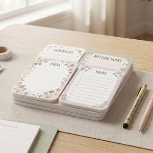 May include: A stack of rectangular paper notepads with rounded corners, featuring printed headers labeled REMINDERS, MEETING NOTES, and IDEAS. The pages are decorated with small floral and heart illustrations in soft pink and brown tones.
