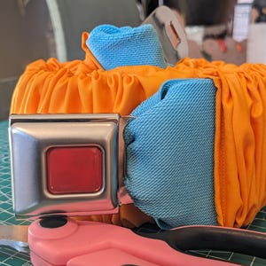 May include: An orange fabric pumpkin with blue fabric accents, secured with a silver seat belt buckle. A pair of pink scissors rests in the foreground on a green cutting mat. The pumpkin is a unique decorative item.