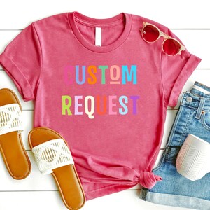 May include: A pink t-shirt with the words "CUSTOM REQUEST" in rainbow colors. The shirt is laid out on a white wooden surface with a pair of white sandals, a pair of sunglasses, and a pair of denim shorts.