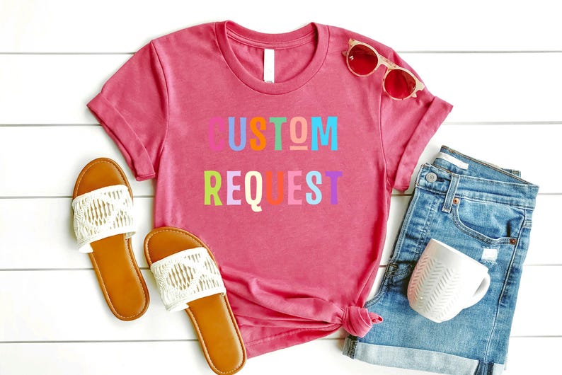 May include: A pink t-shirt with the words "CUSTOM REQUEST" in rainbow colors. The shirt is laid out on a white wooden surface with a pair of white sandals, a pair of sunglasses, and a pair of denim shorts.