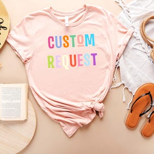 May include: A pink t-shirt with the text "CUSTOM REQUEST" in rainbow colors. The shirt is laid out on a light brown surface with a straw hat, a book, a straw bag, and sandals.