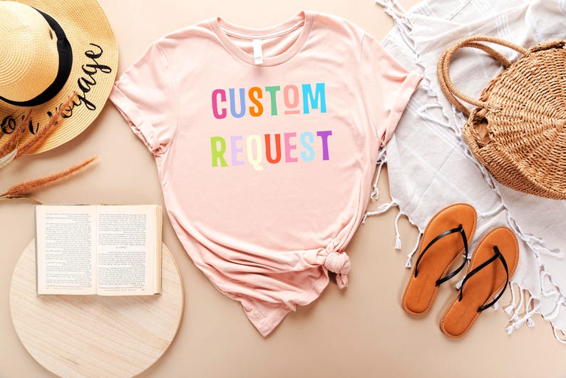 May include: A pink t-shirt with the text "CUSTOM REQUEST" in rainbow colors. The shirt is laid out on a light brown surface with a straw hat, a book, a straw bag, and sandals.