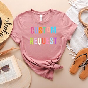 May include: A pink t-shirt with the text "CUSTOM REQUEST" in rainbow colors. The shirt is laid out on a light brown surface with a straw hat, sandals, and a straw bag.