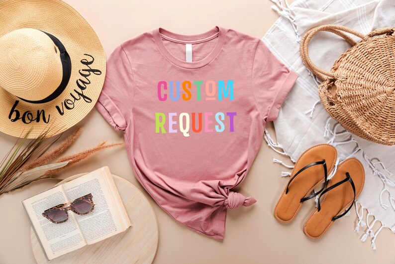 May include: A pink t-shirt with the text "CUSTOM REQUEST" in rainbow colors. The shirt is laid out on a light brown surface with a straw hat, sandals, and a straw bag.