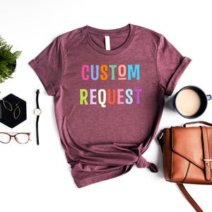 May include: A maroon t-shirt with the text "CUSTOM REQUEST" in rainbow colors. The shirt is tied in a knot at the bottom and is laid out on a white surface.
