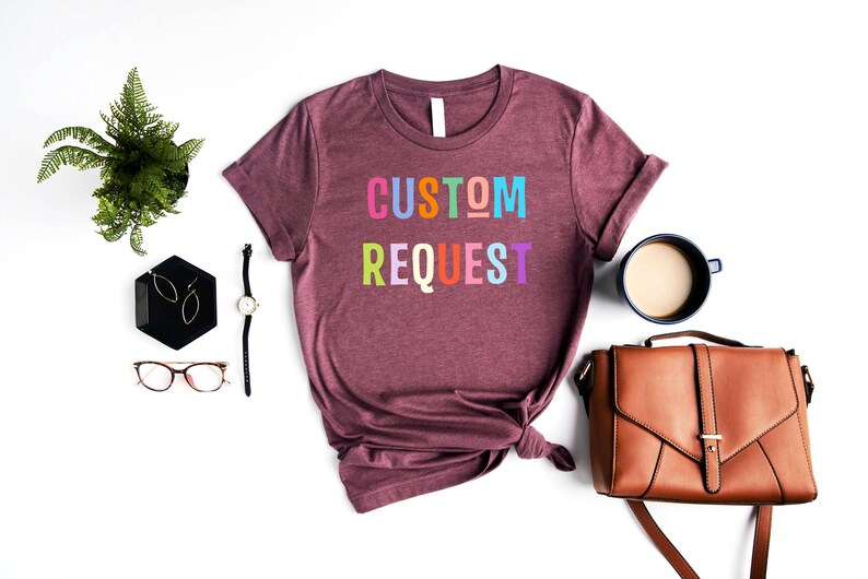 May include: A maroon t-shirt with the text "CUSTOM REQUEST" in rainbow colors. The shirt is tied in a knot at the bottom and is laid out on a white surface.