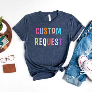 May include: A navy blue t-shirt with the text "CUSTOM REQUEST" in rainbow colors. The shirt is laid flat on a white surface with a pair of jeans and a pair of white sneakers.