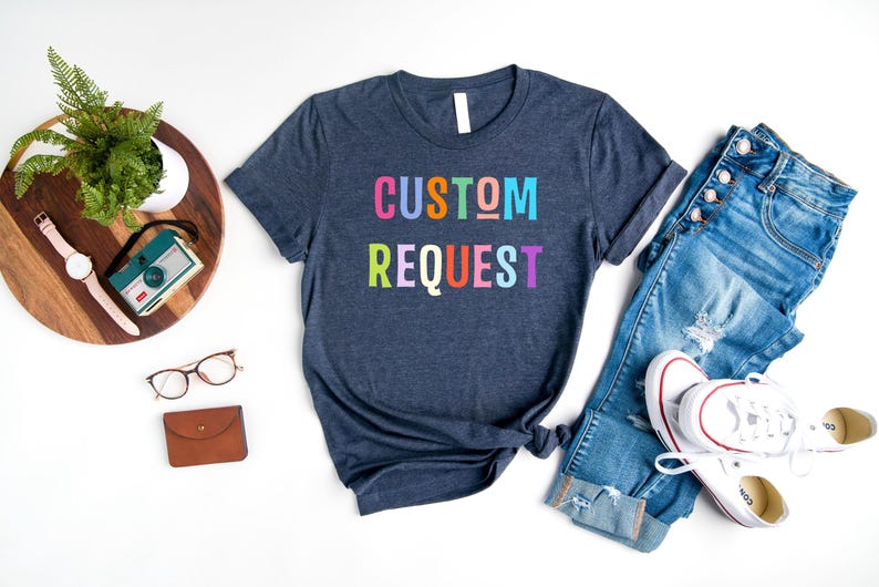 May include: A navy blue t-shirt with the text "CUSTOM REQUEST" in rainbow colors. The shirt is laid flat on a white surface with a pair of jeans and a pair of white sneakers.