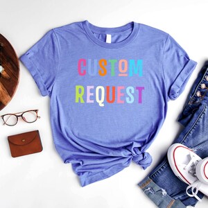 May include: A purple t-shirt with the text "CUSTOM REQUEST" in rainbow colors. The shirt is laid out on a white surface with a pair of jeans, a wallet, and a pair of glasses.
