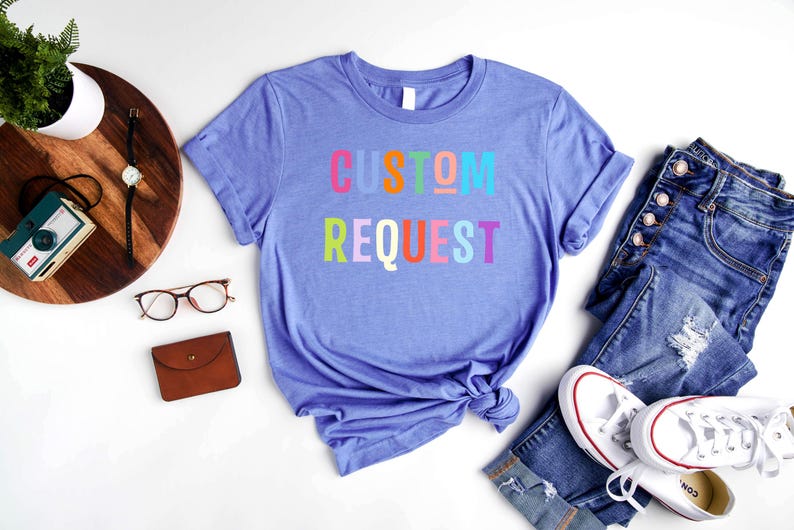 May include: A purple t-shirt with the text "CUSTOM REQUEST" in rainbow colors. The shirt is laid out on a white surface with a pair of jeans, a wallet, and a pair of glasses.