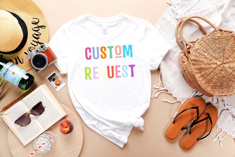 May include: White t-shirt with the text "CUSTOM REQUEST" in rainbow colors. The shirt is laid flat on a light brown surface with various accessories around it, including a straw hat, a camera, a book, a glass of ice water, a peach, and a straw bag.