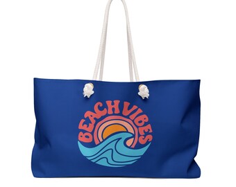 May include: A large, royal blue tote bag with white rope handles. The bag features a circular graphic with the words "BEACH VIBES" in a retro font, along with a stylized sun and ocean wave design in orange, pink, and blue.
