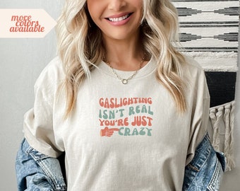 Puede incluir: Una camiseta beige claro con un estampado gráfico que dice "Gaslighting isn't real you're just crazy" en una fuente retro con una mano señalando.