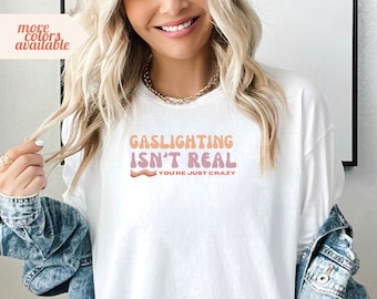 Puede incluir: Camiseta blanca con un gráfico rosa y naranja que dice "Gaslighting Isn't Real You're Just Crazy".