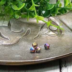 May include: Close-up of four iridescent, faceted beads. The beads are a mix of colors, including blue, purple, and gold. They are resting on a decorative, metallic dish with a leaf design. Green foliage is in the background.