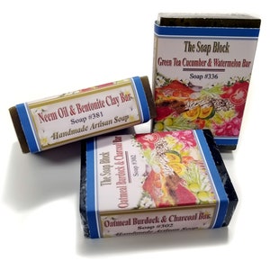 May include: Three bars of handmade artisan soap. The first bar is green and labeled "Neem Oil & Bentonite Clay Bar, Soap #381". The second bar is green and labeled "The Soap Block, Green Tea Cucumber & Watermelon Bar, Soap #336". The third bar is black and labeled "The Soap Block, Oatmeal Burdock & Charcoal Bar, Soap #302".
