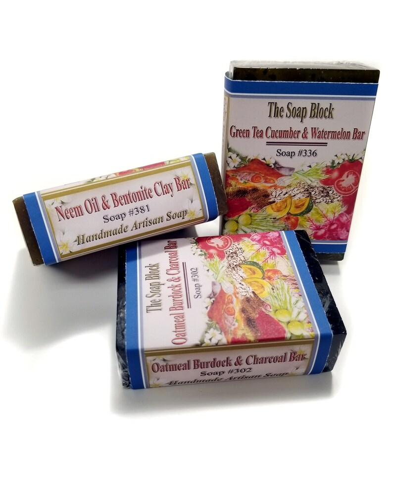 May include: Three bars of handmade artisan soap. The first bar is green and labeled "Neem Oil & Bentonite Clay Bar, Soap #381". The second bar is green and labeled "The Soap Block, Green Tea Cucumber & Watermelon Bar, Soap #336". The third bar is black and labeled "The Soap Block, Oatmeal Burdock & Charcoal Bar, Soap #302".