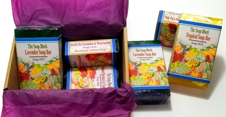 May include: A gift box filled with four bars of handmade soap. The soaps are wrapped in colorful paper with images of fruits and vegetables. The soaps are labeled with the names "The Soap Block Lavender Soap Bar", "Green Tea Cucumber & Watermelon Handmade Artisan Soap", "The Soap Block Lavender Soap Bar", and "The Soap Block Tropical Soap Bar".