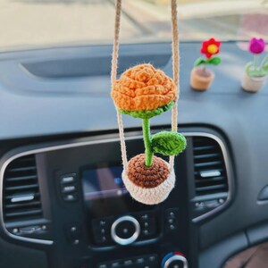 Könnte beinhalten: Eine gehäkelte orangefarbene Blume mit grünen Blättern in einem braunen und weißen Topf, der am Rückspiegel eines Autos hängt.