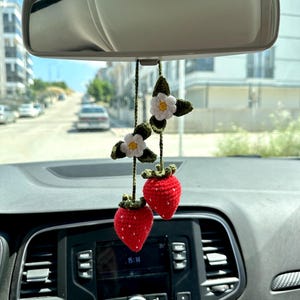 Puede incluir: Una fresa de ganchillo roja y verde colgando de un espejo retrovisor en un coche. La fresa está decorada con flores blancas de ganchillo.