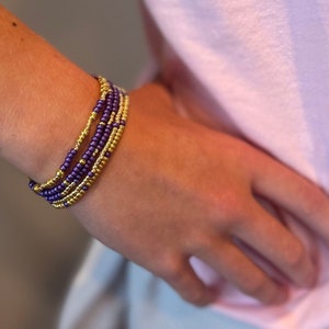 May include: A stack of purple and gold beaded bracelets on a person's wrist.