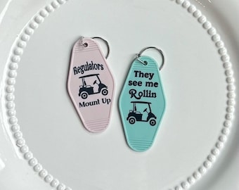 May include: Two pink and blue keychains shaped like motel room keys. The pink keychain has a golf cart and the text "Regulators Mount Up". The blue keychain has a golf cart and the text "They see me Rollin".