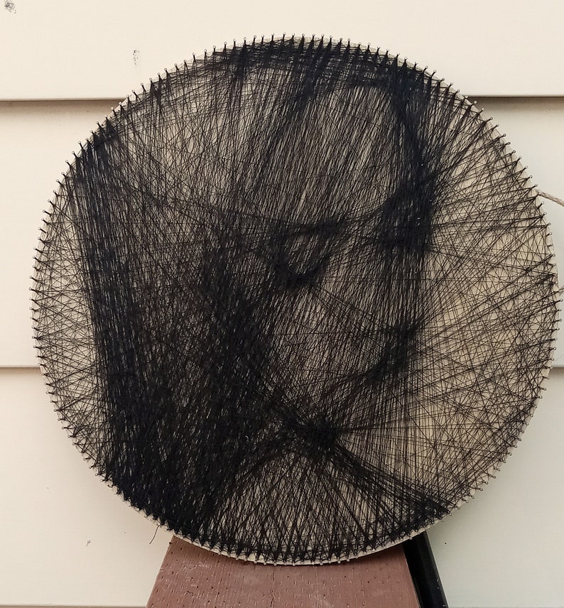 Może przedstawiać: Okrągły kawałek drewna z czarną nicią owiniętą wok&oacute;ł gwoździ, tworząc wz&oacute;r twarzy.