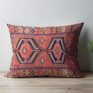 以下が含まれることがあります： ヴィンテージラグのデザインが施された長方形のクッション。赤、ネイビーブルー、黒の色合いで中央に幾何学模様があり、詳細なボーダーで縁取られています。伝統的で手作りの美学を連想させるデザインです。