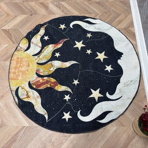 Puede incluir: Una alfombra redonda de color negro y blanco con un diseño de sol y luna. El sol es amarillo y naranja con rayos rojos y blancos. La luna es blanca con una cara y está rodeada de estrellas. La alfombra está sobre un suelo de madera.