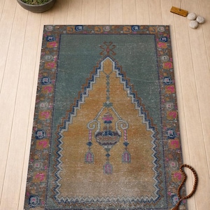 Op de afbeelding: Een rechthoekig vloerkleed in vintage-stijl met een centraal ontwerp in tinten goud en blauwgroen. Het vloerkleed heeft een gedetailleerde rand met roze en blauwe accenten. Het ontwerp omvat een centrale, gestileerde boogvorm. Het vloerkleed ligt op een lichte houten vloer.
