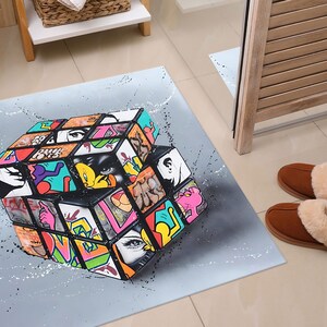 Può includere: Un tappeto grigio e bianco con un design a cubo 3D colorato. Il cubo è composto da quadrati più piccoli con colori e motivi diversi, tra cui lettere e immagini in stile graffiti. Il tappeto è perfetto per aggiungere un tocco di colore e personalità a qualsiasi stanza.