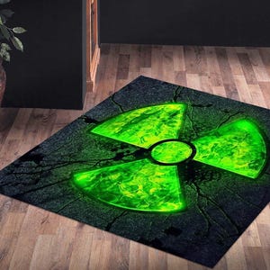 May include: A rectangular rug featuring a glowing green radiation symbol on a dark, cracked background. The symbol is a vibrant green with a black outline, set against a textured, dark gray surface. The rug's design evokes a sense of danger and energy.