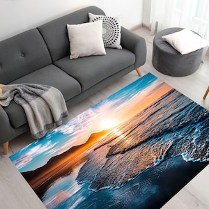 Könnte beinhalten: Ein Teppich mit einer Strandszene, die einen Sonnenuntergang über dem Meer mit Wellen zeigt, die an das Ufer krachen. Der Teppich hat eine blaue, orange und weiße Farbgebung.