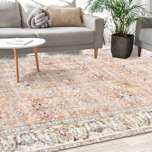 Puede incluir: Una alfombra de área de color melocotón claro y crema con un patrón floral vintage. La alfombra tiene un borde blanco con un diseño floral detallado. La alfombra está colocada sobre un suelo de madera clara en un salón.