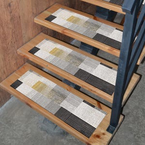 Könnte beinhalten: Holztreppe mit Stufenmatten mit einem geometrischen Muster in Schwarz, Weiß und Gold. Die Matten weisen eine Mischung aus Linien und Punkten auf, die ein modernes Design schaffen. Die Treppe hat ein Metallgeländer.