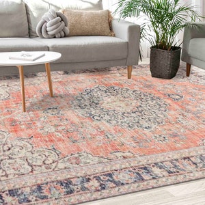 Puede incluir: Una alfombra de estilo vintage con un aspecto descolorido y desgastado. La alfombra presenta un medallón central en tonos azules, crema y rosa, rodeado de un borde de intrincados patrones florales. La alfombra está colocada sobre un suelo de madera clara.