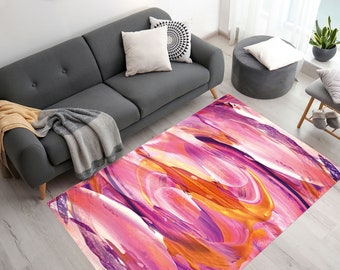 Op de afbeelding: Abstract vloerkleed met roze, oranje en paarse verfstreken. Het kleed ligt op een lichte houten vloer in een woonkamer.
