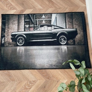 Puede incluir: Una alfombra negra y gris con un coche vintage impreso en ella. El coche es un coche americano clásico de músculo con un cuerpo negro y un interior gris. La alfombra es rectangular y tiene un borde negro.
