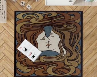 Puede incluir: Una alfombra cuadrada con un diseño Art Nouveau que presenta dos caras besándose, con cabello castaño fluido y un fondo azul. La alfombra tiene un borde negro y un libro encima.